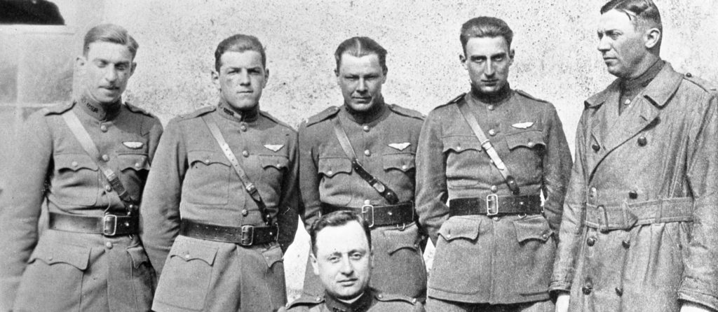 A photo of five men in uniform standing, in front of a sixth man, apparently seated.