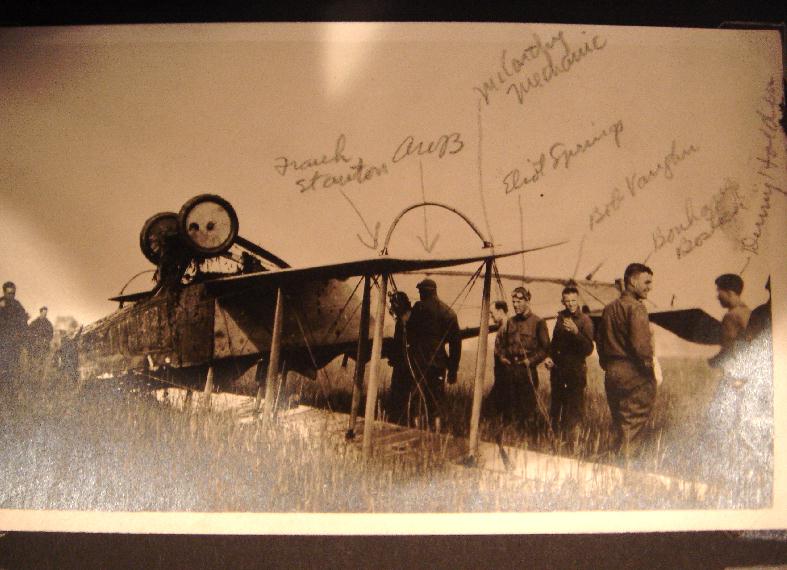 Photo of an upside down plane in a field, with a number of men inspecting it. Names have been written in identifying, among others, Springs, Vaughn, and Bostick.