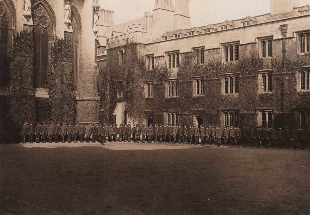 Photo of a long line of uniformed men standing in front of the towering, ivy covered walls of gothic buildings; they are in the northeast corner of the front quadrangle of Exeter College, Oxford.