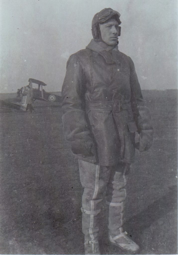 A man in flying clothes and leather flying helmet in the foreground, with a Sopwith Camel in the background.