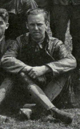 Photo of a man sitting on the ground with legs crossed; it is evidently cropped from a group photo.