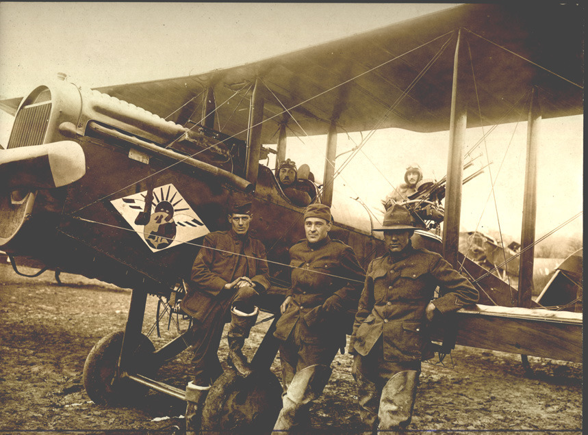 A photo of a DH-4 with Devery in the cockpit and Henry Irving Jenks acting as gunner. There are also three men in the foreground leaning against the plane.
