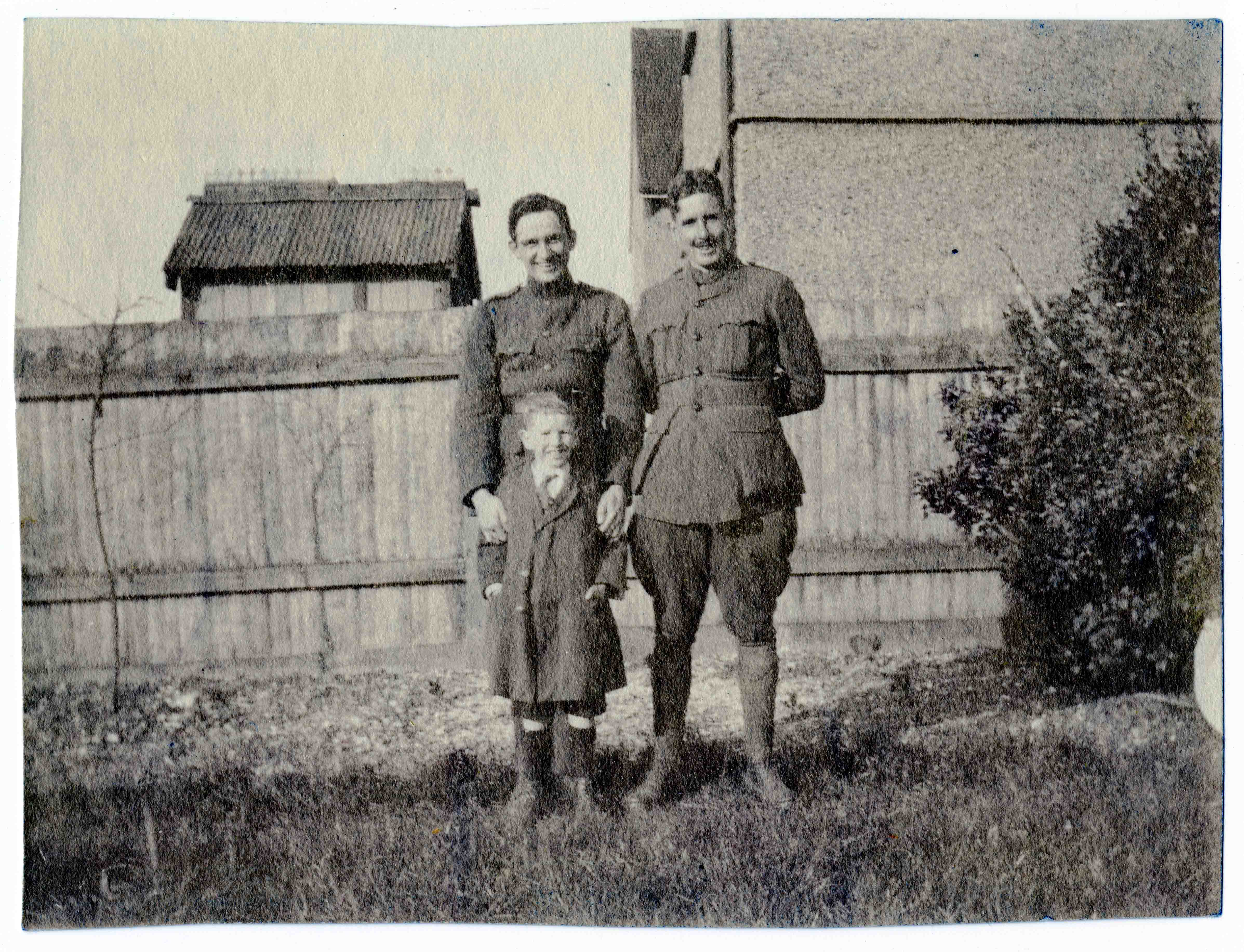 A photo of two men in uniform, but without pilots' wings, outdoors, apparently in the backyard of a house, standing in front of a boy, about seven years old.