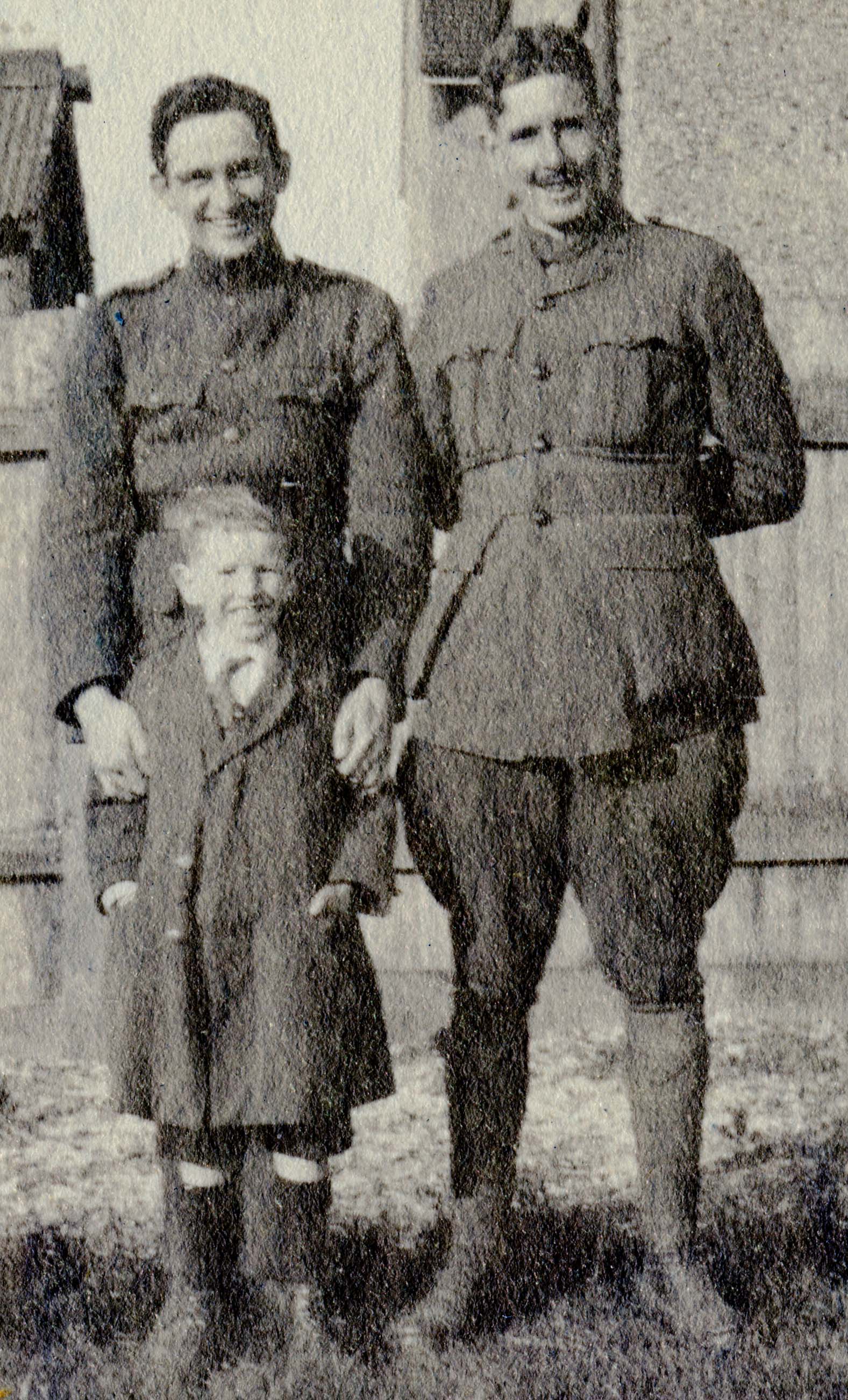 A photo of two men in uniform, but without pilots' wings, outdoors, apparently in the backyard of a house, standing in front of a boy, about seven years old.