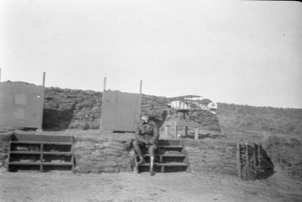 Man in uniform sitting on steps against a background of sandbags and targets, with a mock airplane in the background.