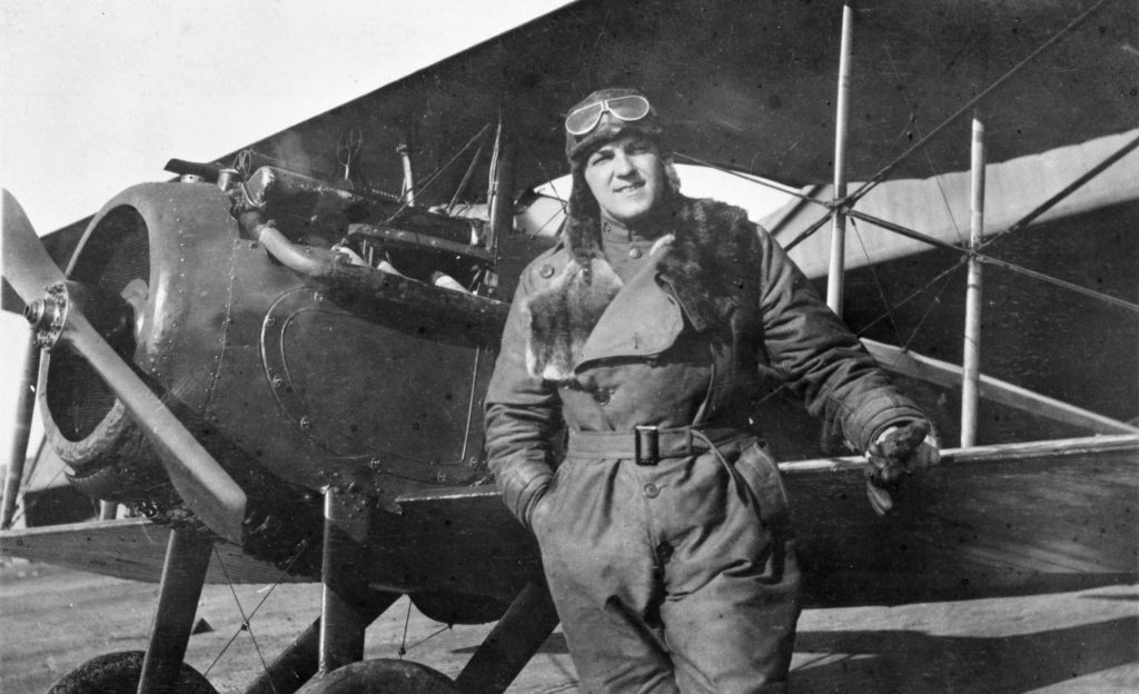 A pilot in flying clothes and helmet with goggles, in front of a Spad VII.