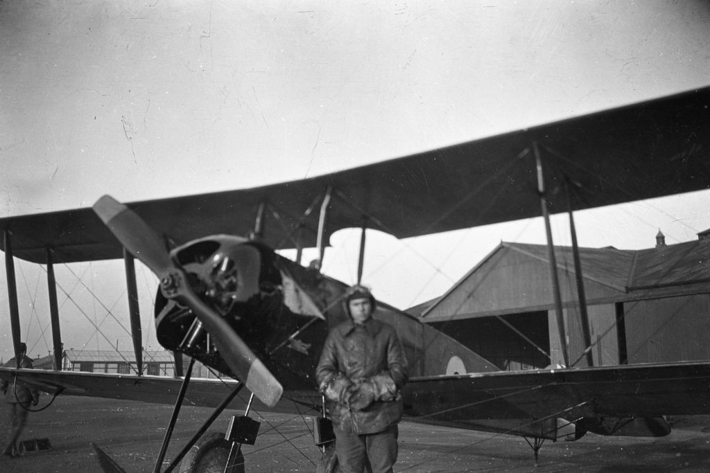 Black and white photo of a plane (an Avro) in front of a large hangar, with a man in leather flying clothes and helmet standing in front of the plane.