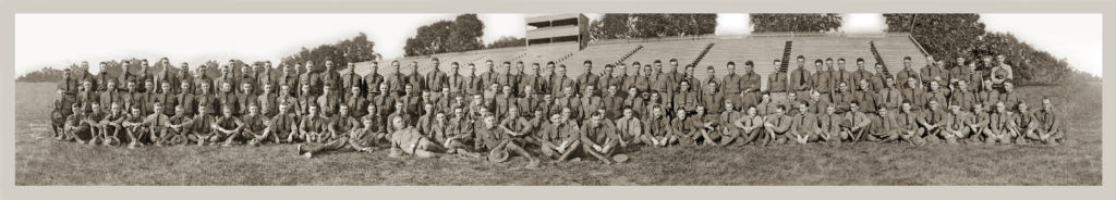 A wide angle picture of approximately 120 uniformed men in four rows in front of outdoor bleachers.