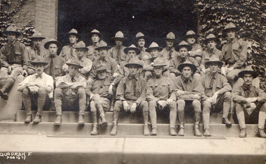 Photo of twenty-four men wearing campaign hats seated in three rows on the steps of a building with ivy covered walls. Written in the lower left corner is the notation "Squadron F. august 10 '17."