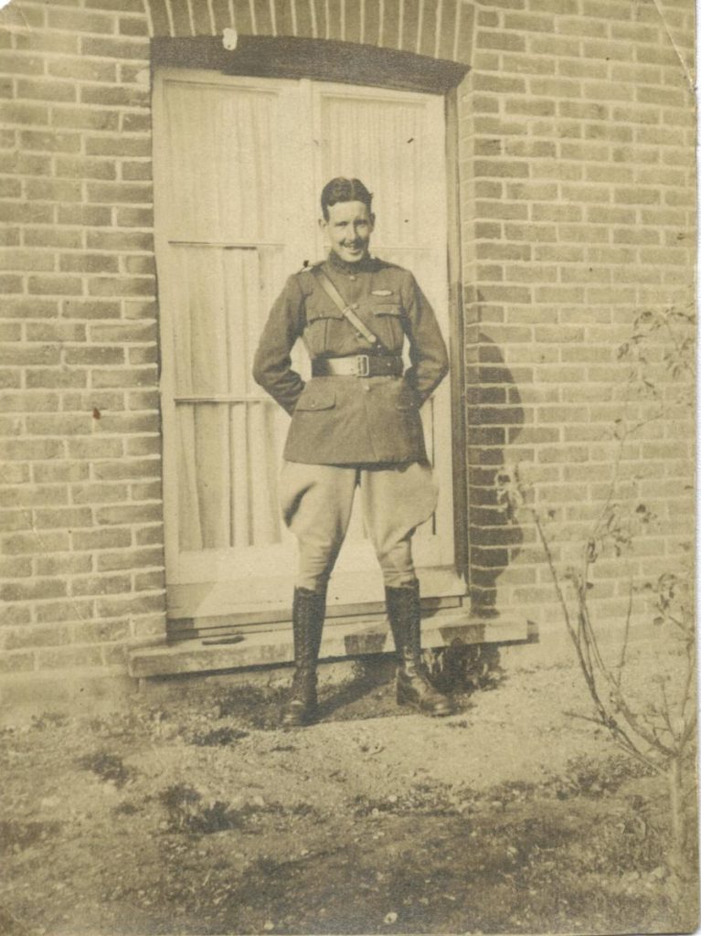 Photo of a smiling Curtis in uniform with his pilots wings above his left breast pocket; he is standing before a tall window outside a brick house.