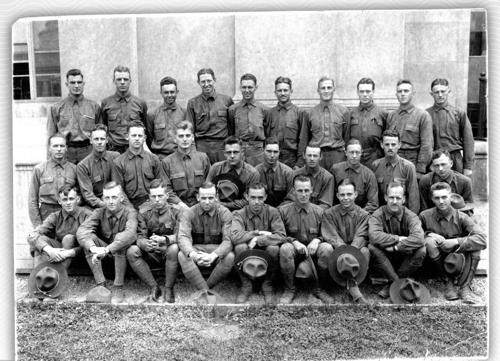 Photo of nineteen men in three rows, all dressed similarly in long sleeved Oxford shirts without ties. Many of the men seated in the front row are holding campaign hats, but all are bare-headed.