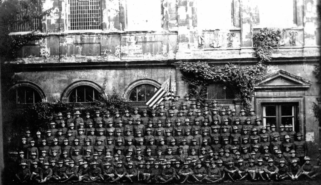 Large group photo of about 180 men in uniform ranged in about 10 rows before an American flag and ivy-covered walls.